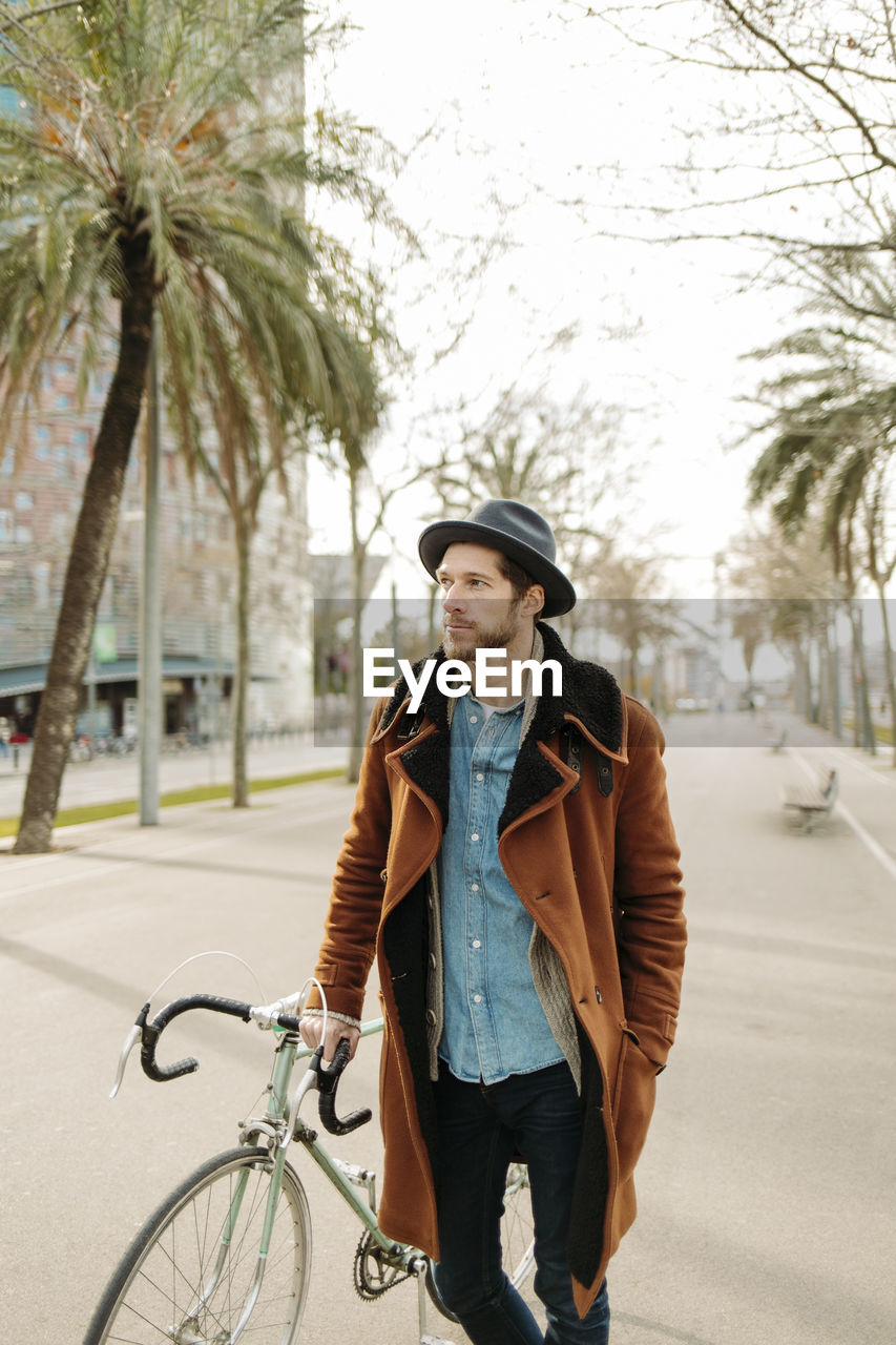 Hipster man wearing hat standing with bicycle in city while looking away