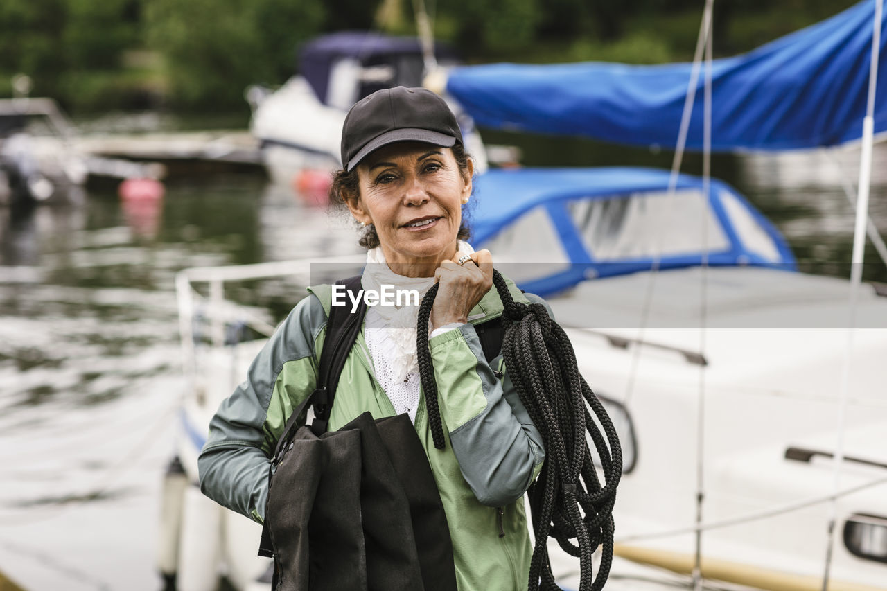 Portrait of confident female instructor holding rope and life jackets against yacht during boat master course