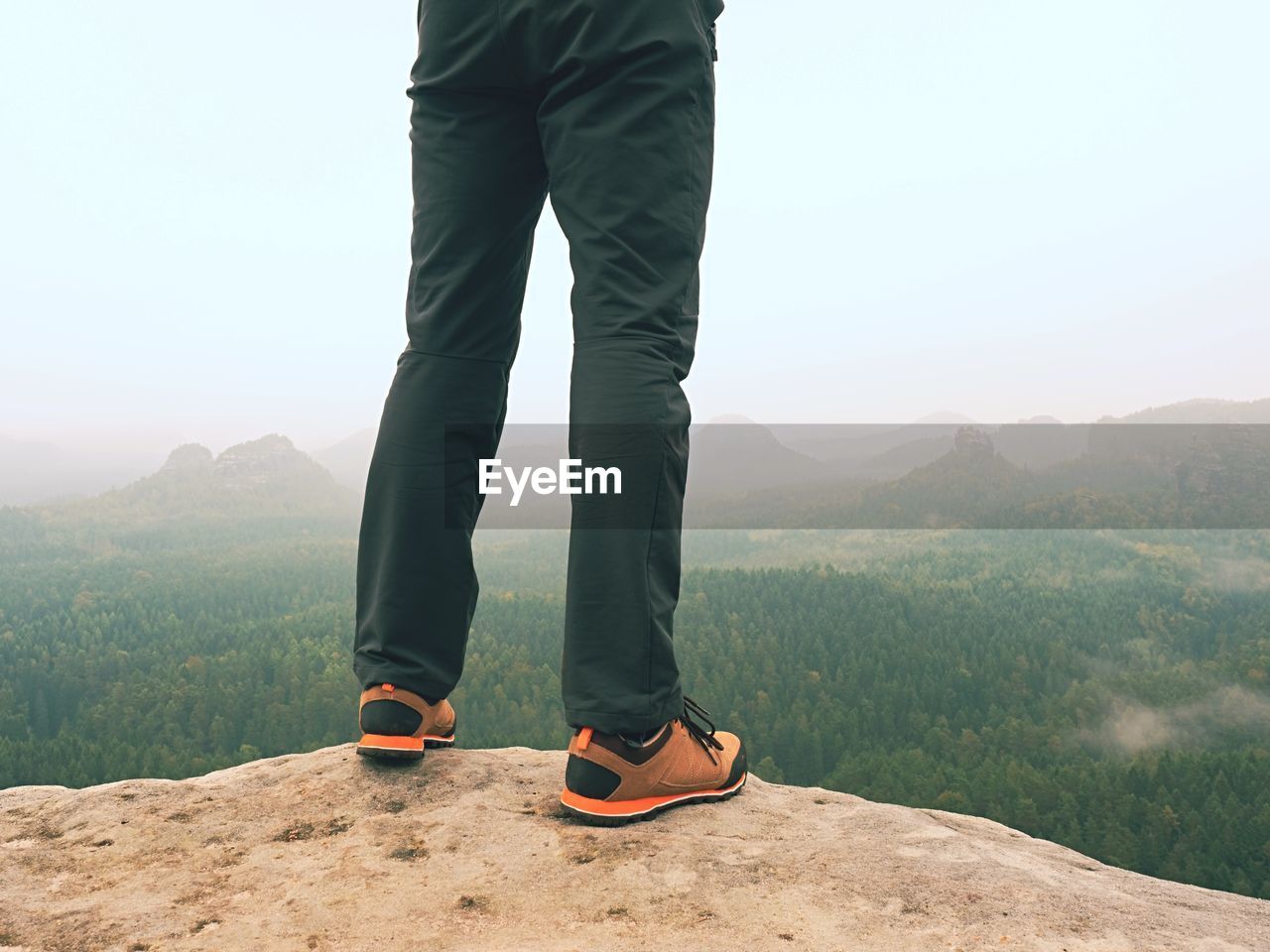 Male legs in dark hiking trousers and leather trekking shoes on peak of rock above misty valley