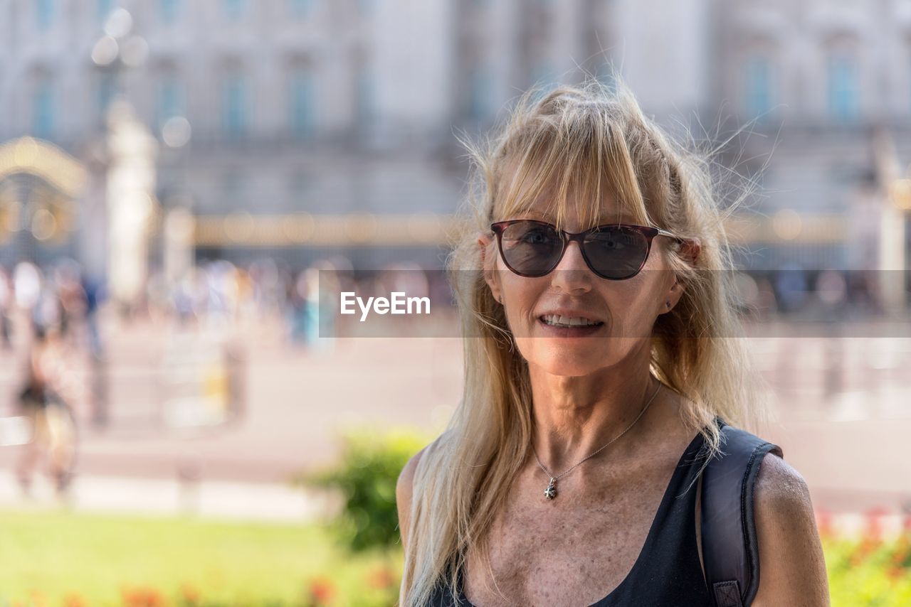 Mature blonde woman with glasses looking at the camera. behind out of focus buckingham palace.