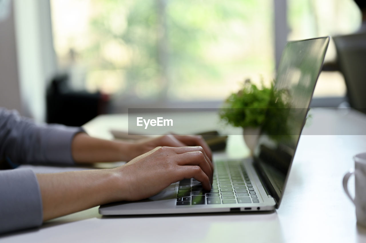 computer, wireless technology, technology, laptop, adult, one person, table, using laptop, communication, business, hand, furniture, indoors, writing, working, desk, office, internet, typing, day, computer network, sitting, corporate business, businessman, computer equipment, convenience, window, young adult, business finance and industry, close-up, selective focus, portability, occupation, businesswoman, home interior, businesswear, person, women, using computer, focus on foreground, side view, lifestyles, finance