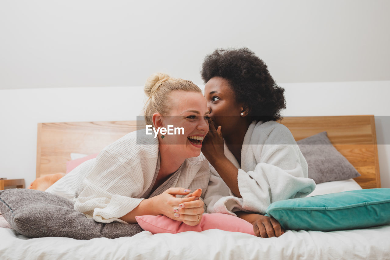 Woman whispering friend while sitting on bed