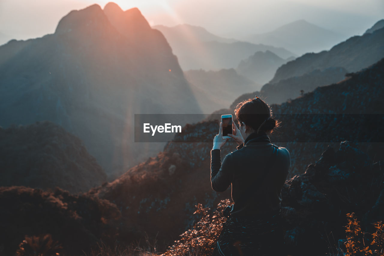 Rear view of woman photographing mountains through mobile phone during sunset