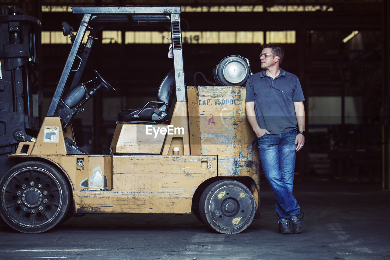 Full length of worker standing by forklift in industry