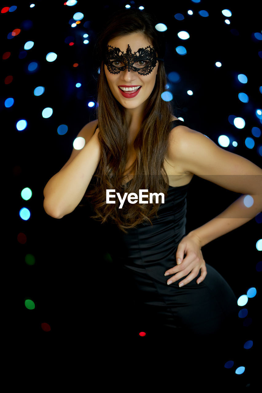 Portrait of beautiful young woman wearing mask standing against illuminated lights