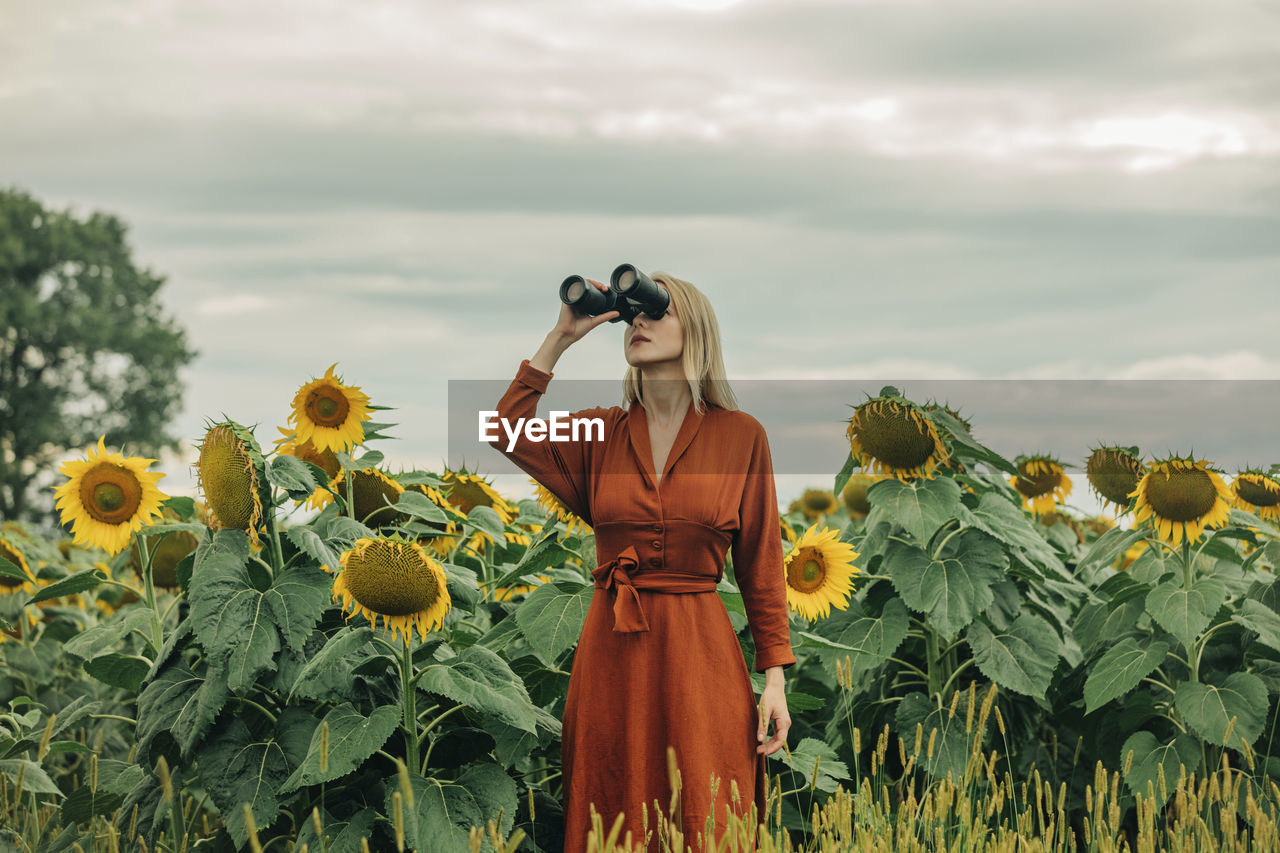 Woman in red dress looking through binoculars at sunflower field