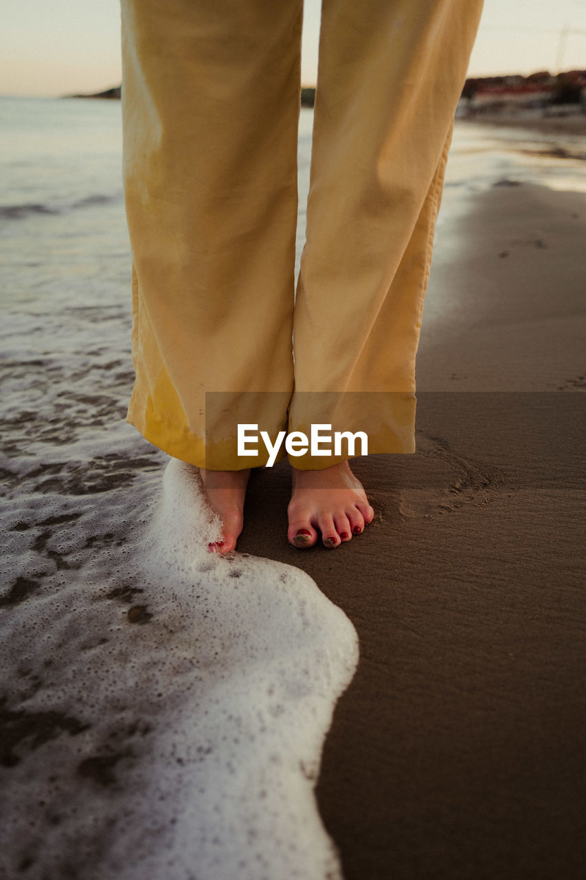 Legs of crop unrecognizable barefoot female standing in foam of wavy ocean in sunset