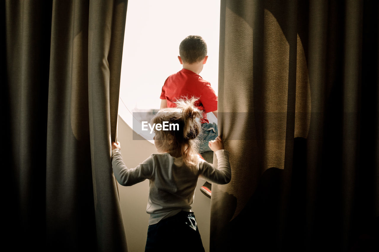 Rear view of girl holding curtain while brother sitting on window in hotel room