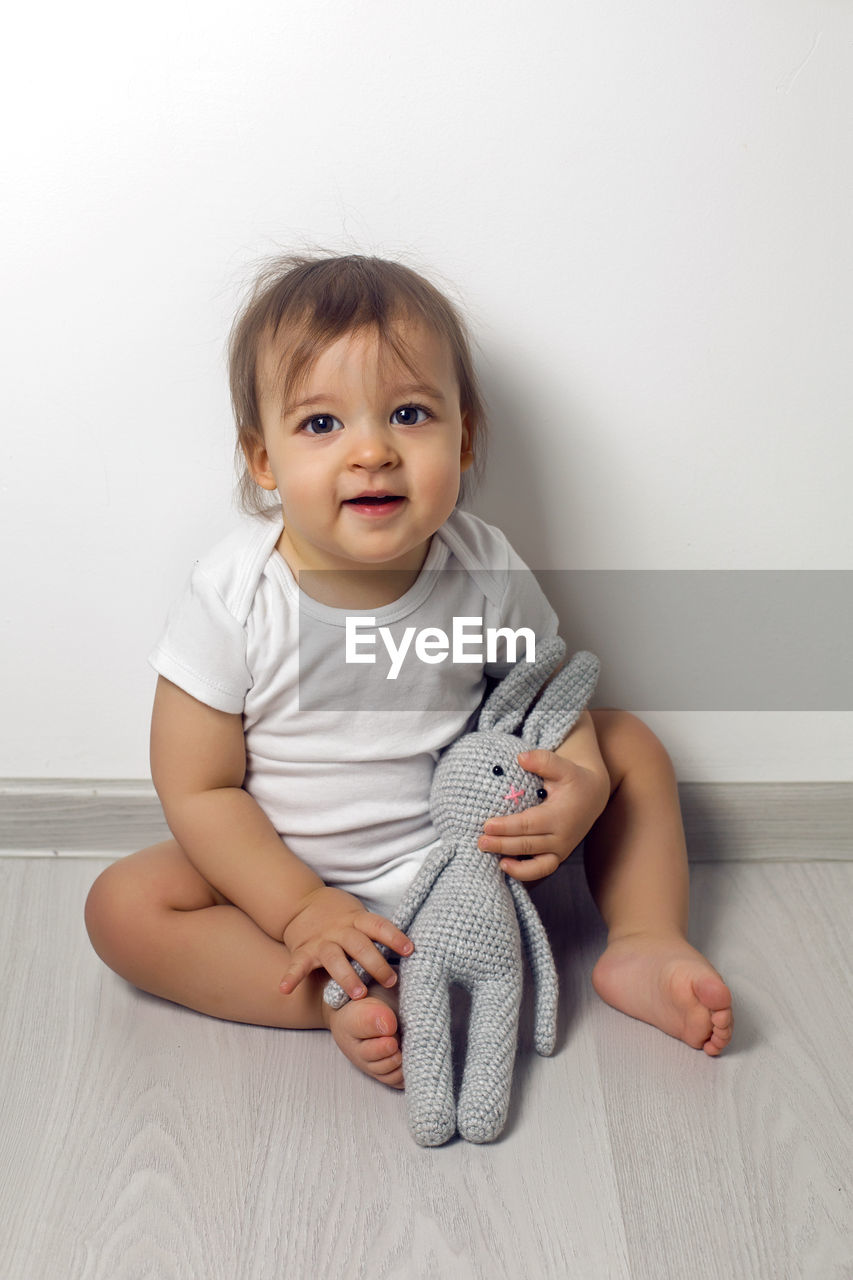 Baby boy in white clothes sitting on the floor with a gray knitted rabbit at the white wall 