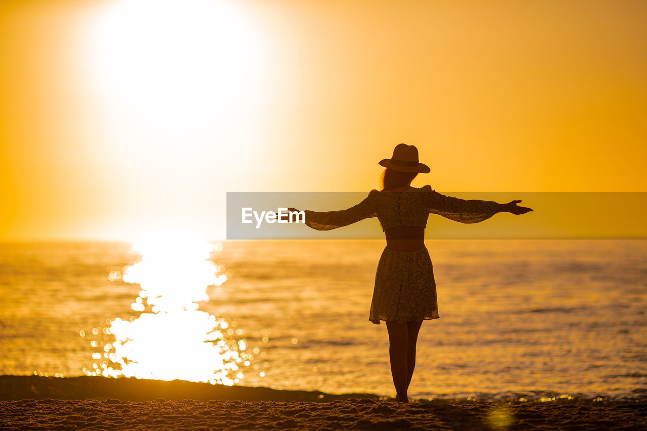 silhouette woman standing at beach against orange sky
