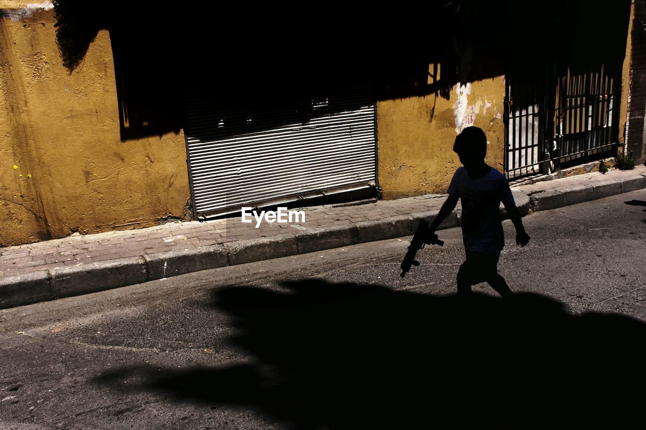 Silhouette boy with toy gun walking on road during sunny day