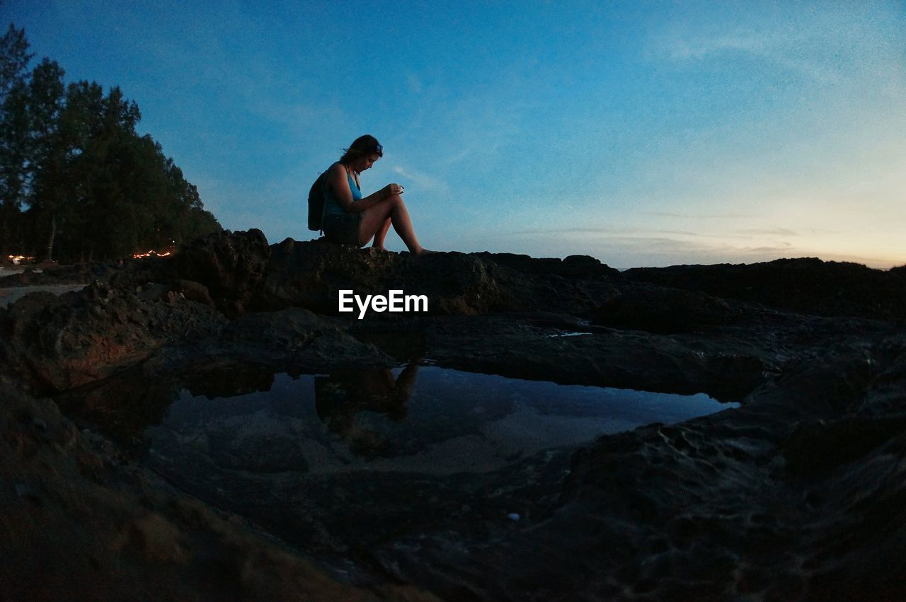 Woman sitting on rock formation at beach against sky during sunset