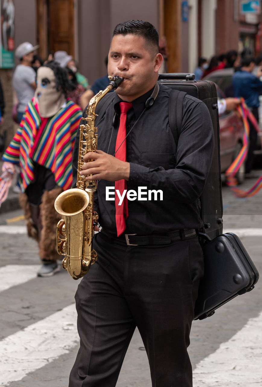 musical instrument, music, city, saxophone, arts culture and entertainment, men, person, street, musician, clothing, adult, architecture, holding, street musician, woodwind instrument, street artist, performance, road, wind instrument, outdoors, young adult, standing, day, saxophonist, focus on foreground, city life