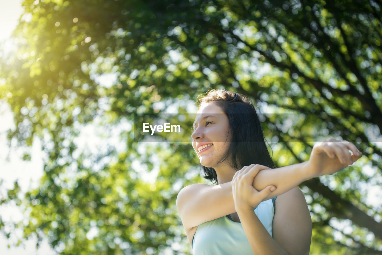 Low angle view of smiling teenage girl looking away while exercising against trees at park