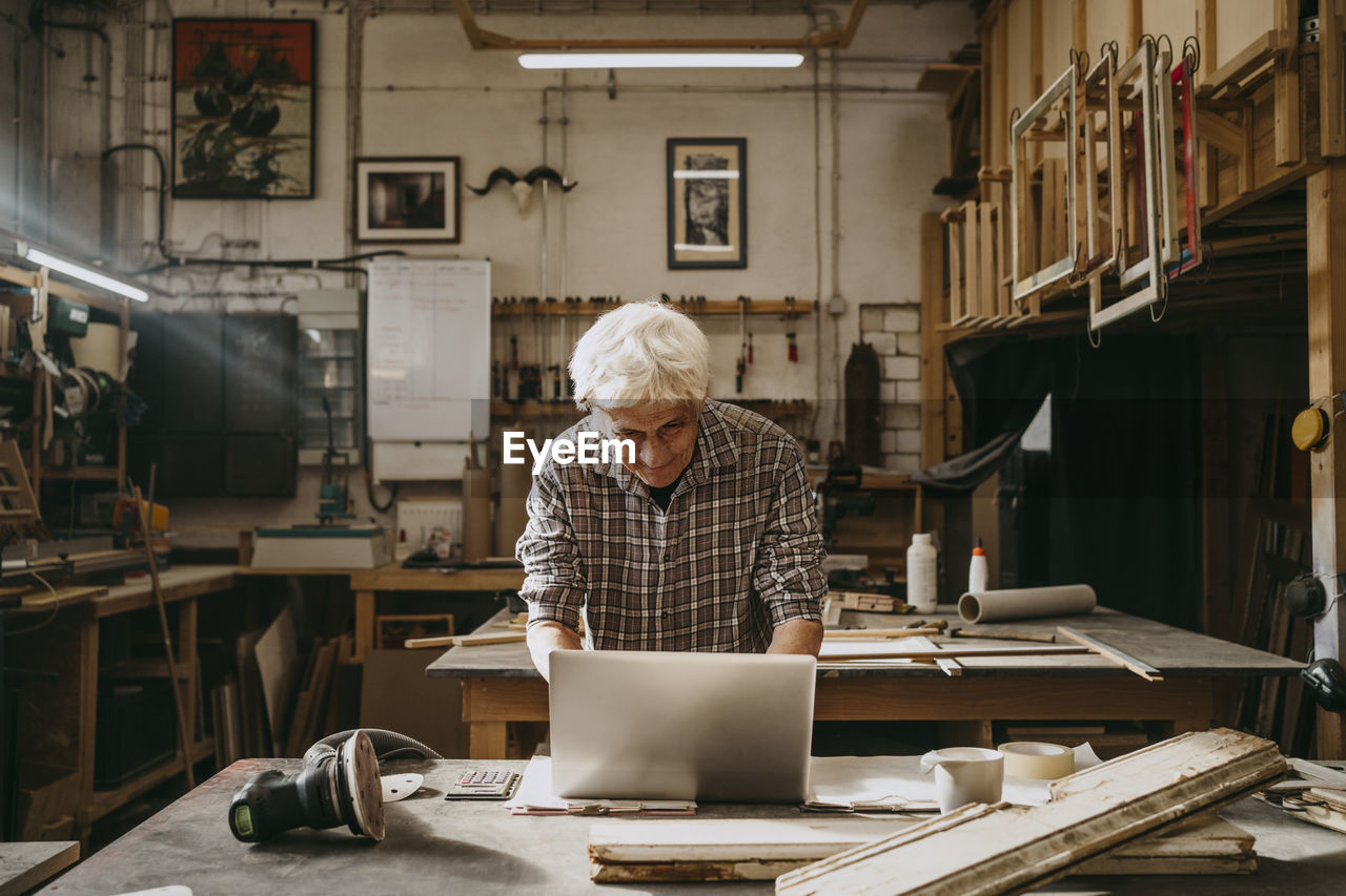 Senior male carpenter working on laptop at repair shop