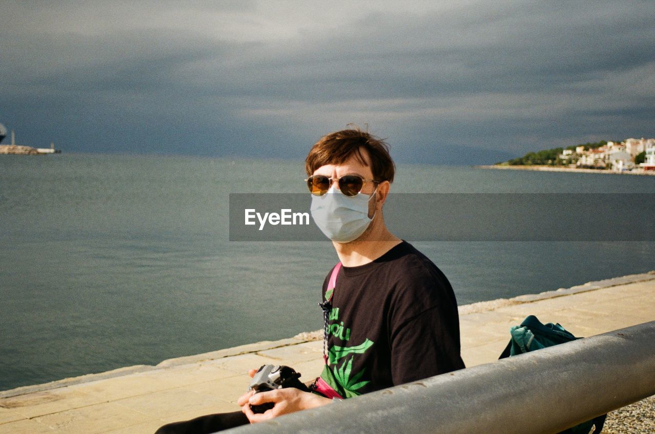 Portrait of man wearing mask and sunglasses sitting against sea