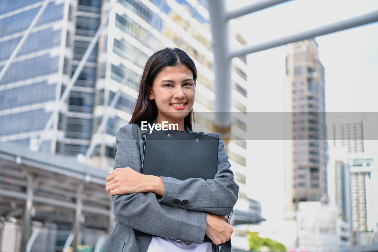 Portrait of businesswoman with file standing against buildings in city