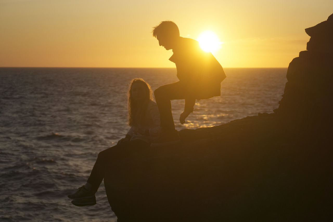 SILHOUETTE MAN SITTING ON BEACH AGAINST SUNSET SKY