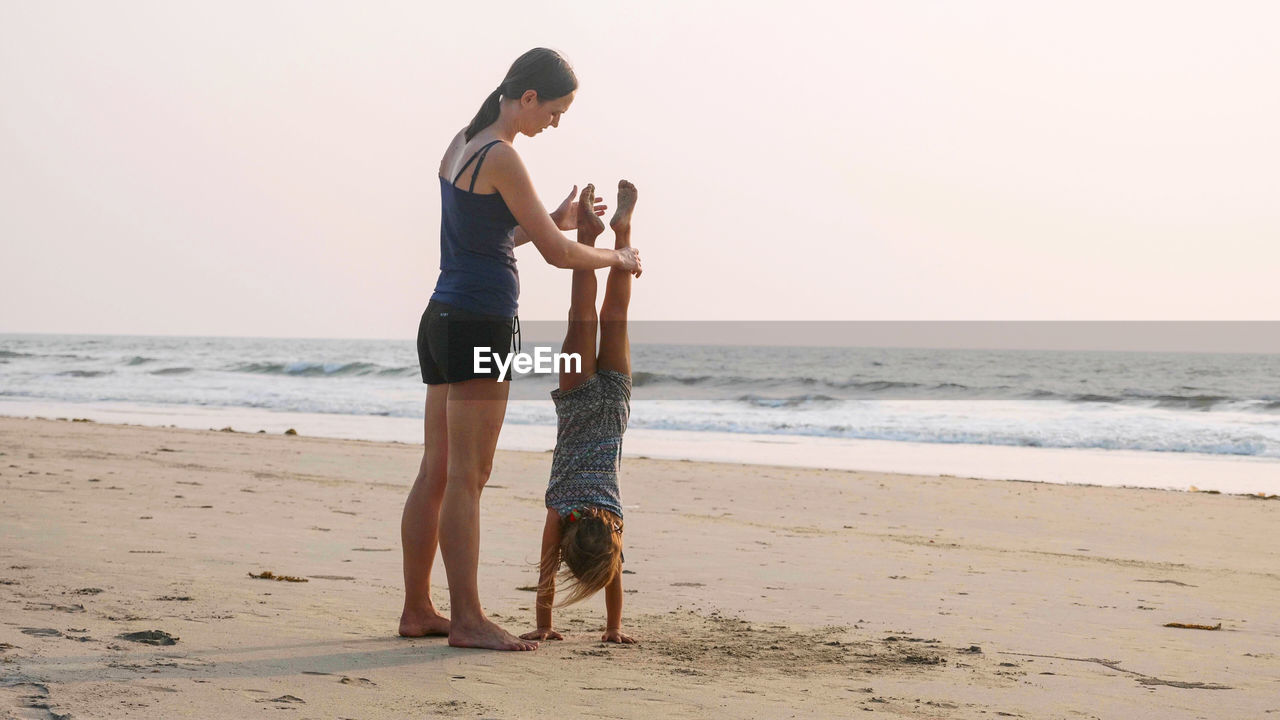 Full length of mother helping daughter in doing handstand on beach against clear sky