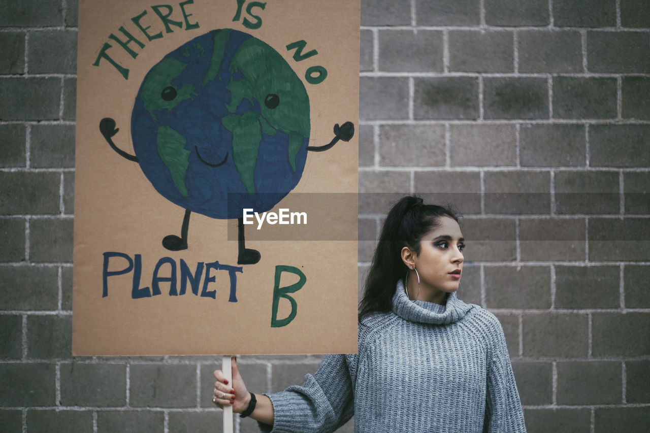 Female activist looking away while holding planet earth poster against wall