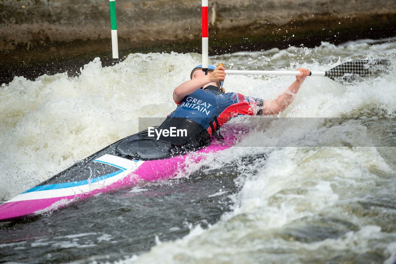 Gb canoe slalom athlete ducking under slalom pole while negotiating upstream gate in white water.