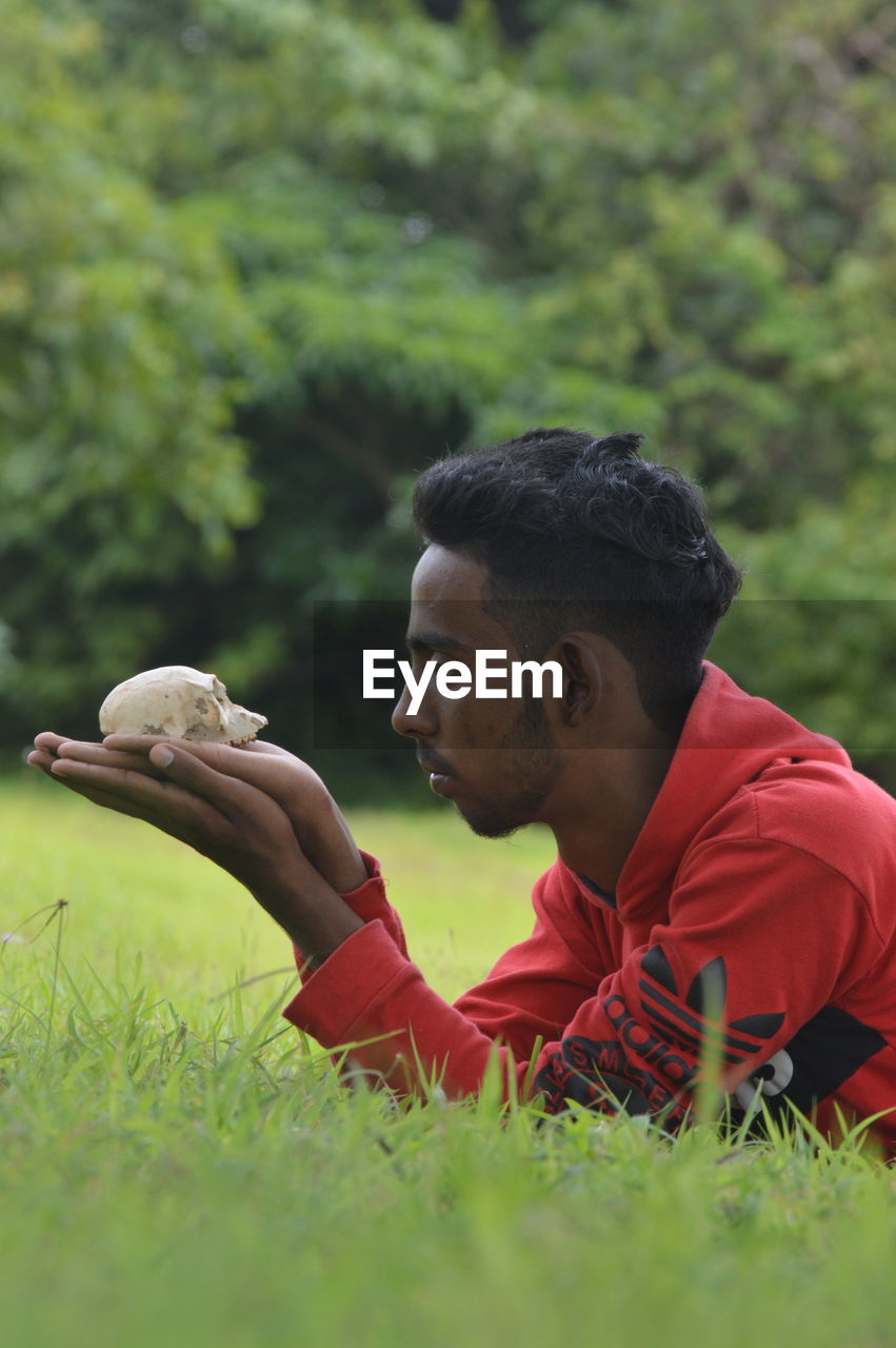 Side view of young man holding rock while lying on grassy land in forest