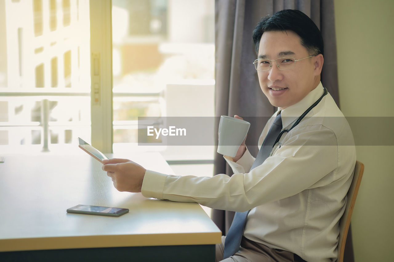 Portrait of smiling businessman drinking coffee while sitting with digital tablet and mobile phone at table in office