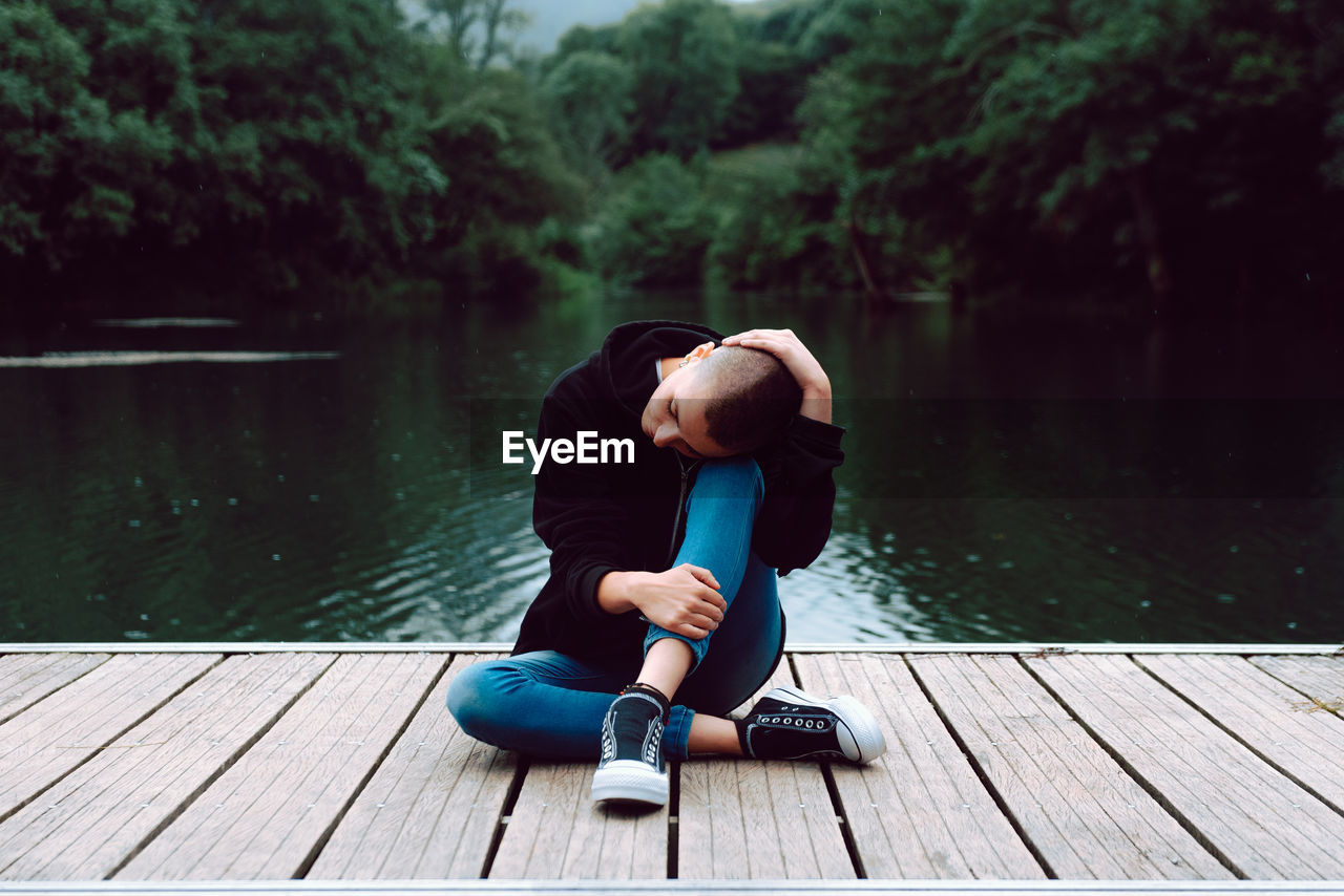 Unique female hipster with short hairstyle in casual clothing sitting with crossed legs on wooden pier of pond with green trees on blurred background