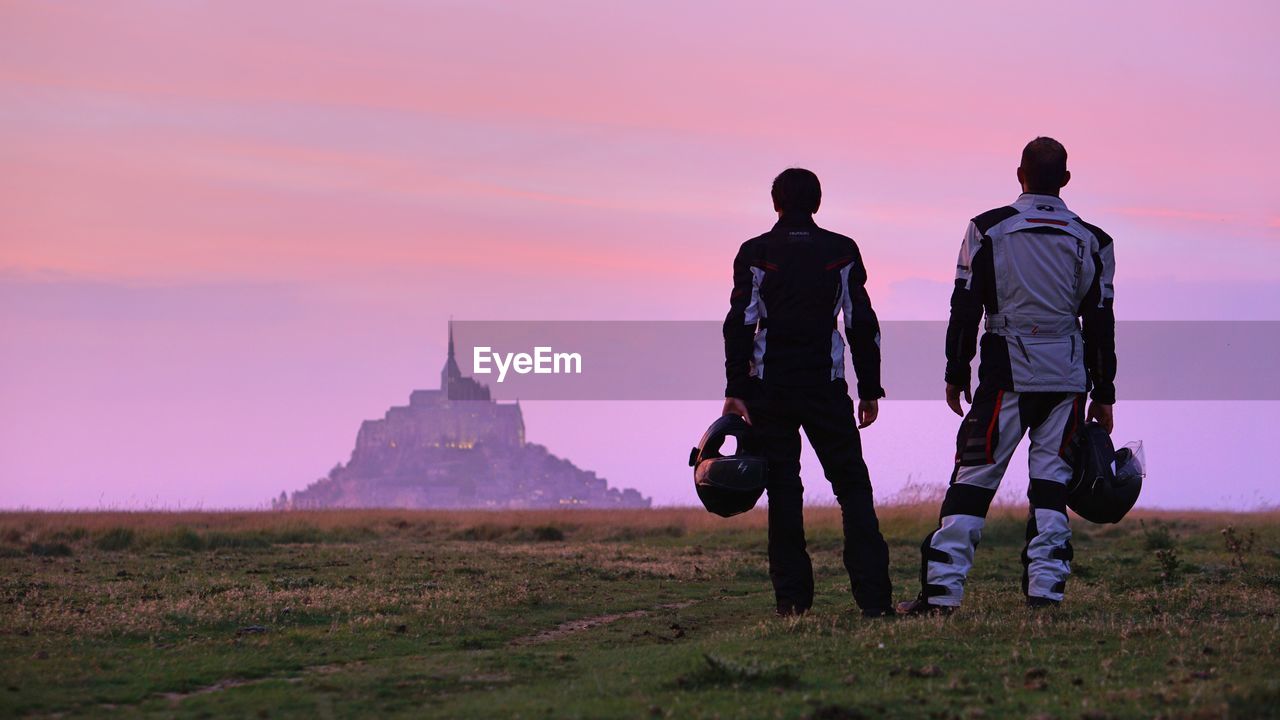 REAR VIEW OF MEN STANDING ON FIELD AGAINST SUNSET SKY