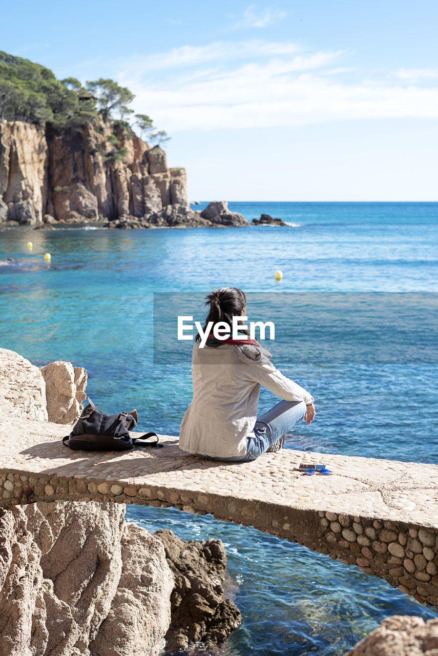 Rear view of a woman relaxing in front of blue sea, sitting on bridge