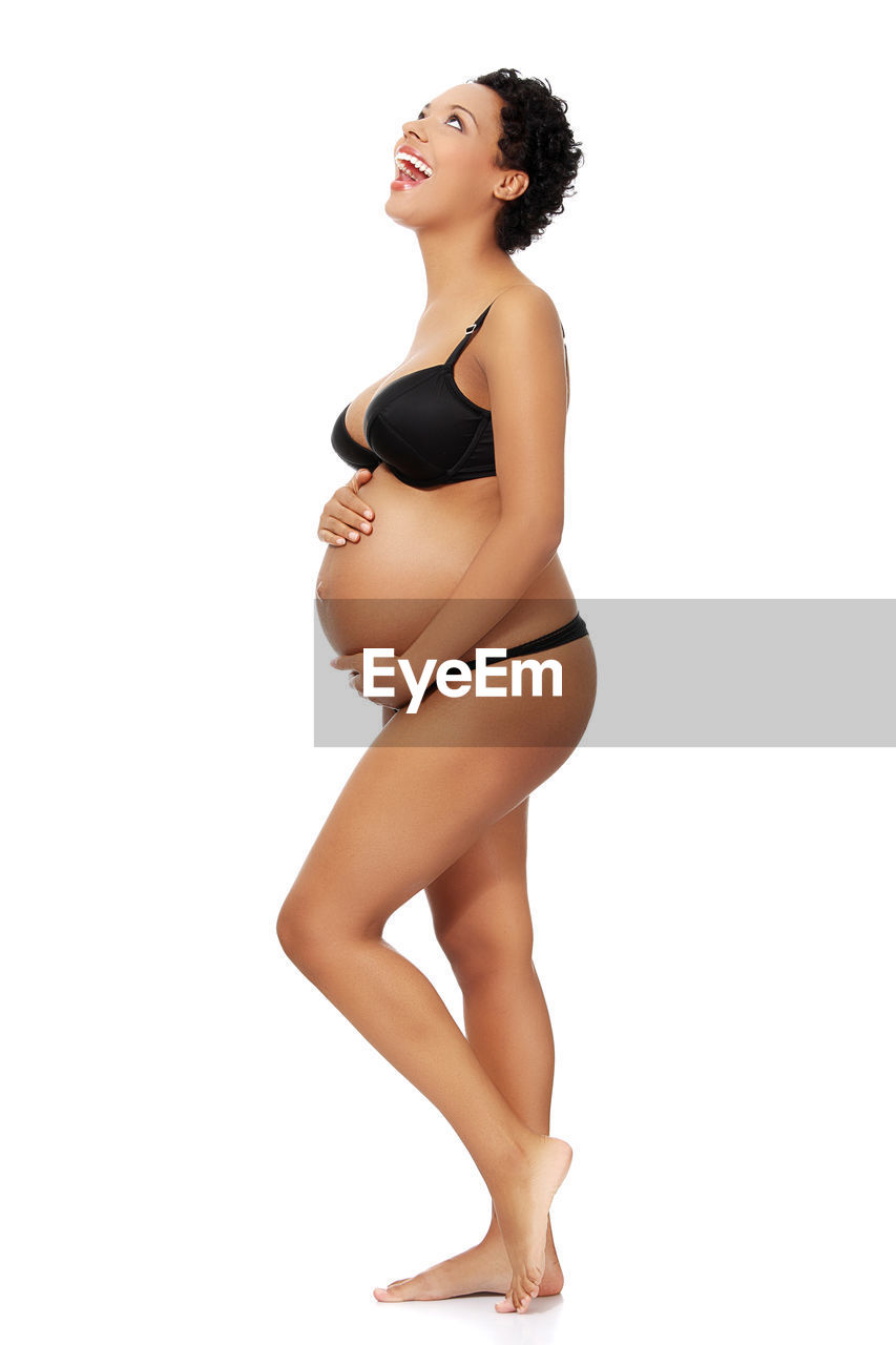 Smiling pregnant woman touching abdomen while standing against white background