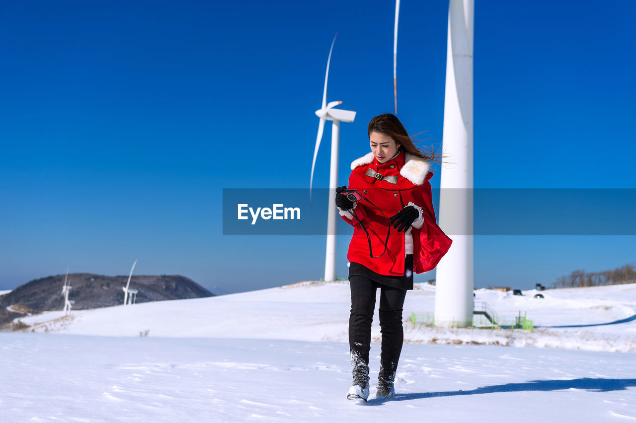 Woman holding camera while walking on snow covered road against wind turbines and clear blue sky