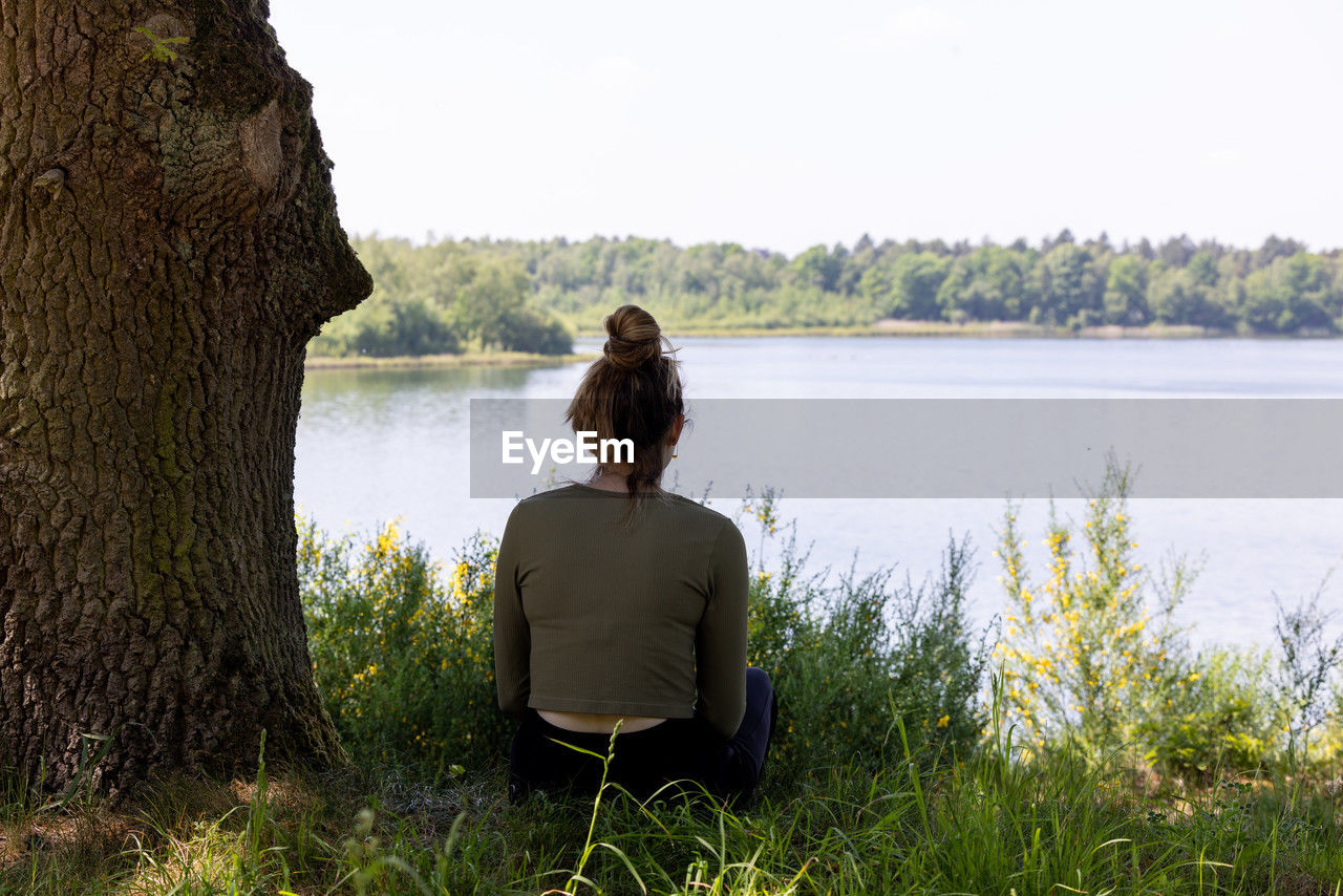 sitting, water, rear view, one person, adult, nature, tranquility, lake, plant, relaxation, women, leisure activity, tree, beauty in nature, hairstyle, tranquil scene, day, vacation, trip, solitude, sky, contemplation, holiday, looking at view, lifestyles, casual clothing, grass, outdoors, long hair, young adult, scenics - nature, three quarter length, land, copy space, looking, environment, person, beach, non-urban scene, simple living, full length, idyllic, brown hair, landscape, clear sky, shore