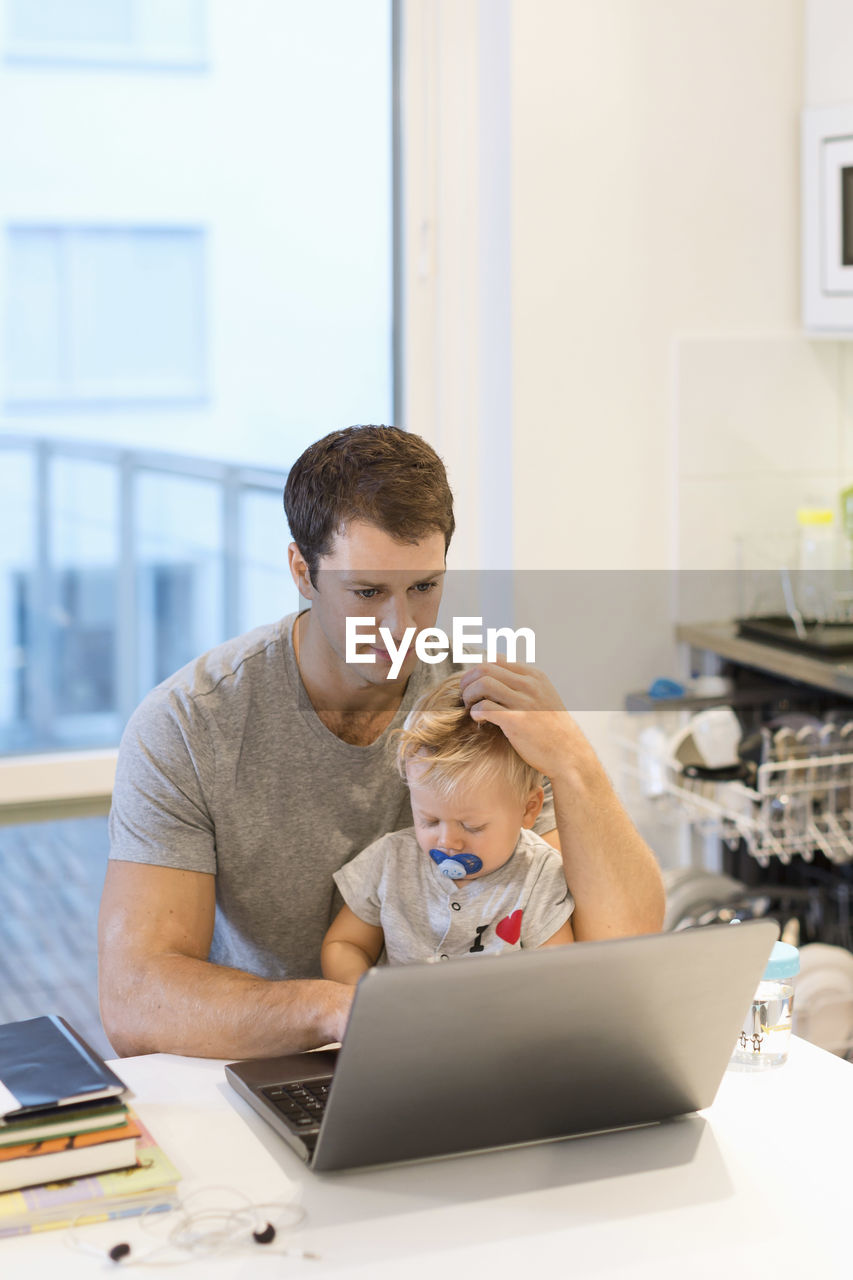 Baby boy sitting with father using laptop at table in house