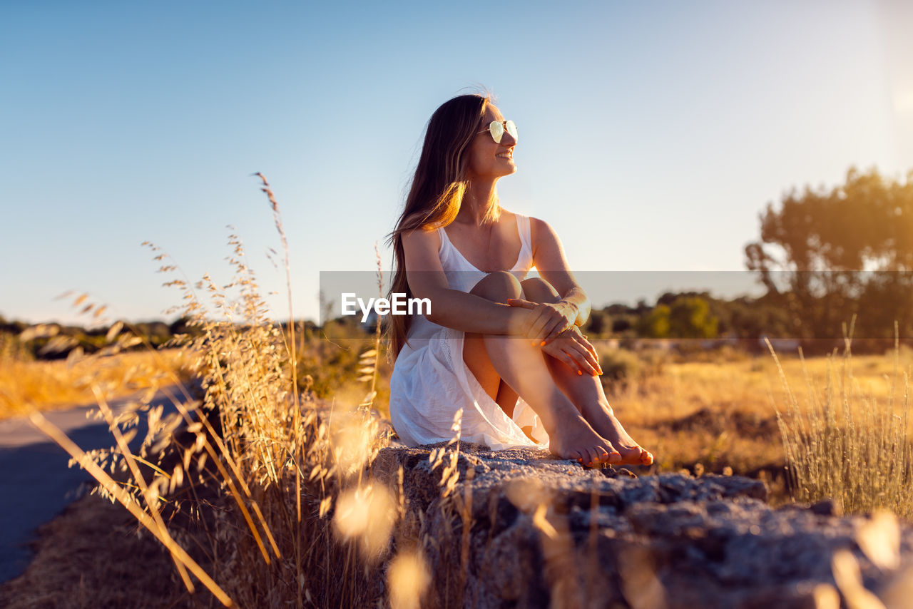 Smiling woman looking away while sitting on rock against sky