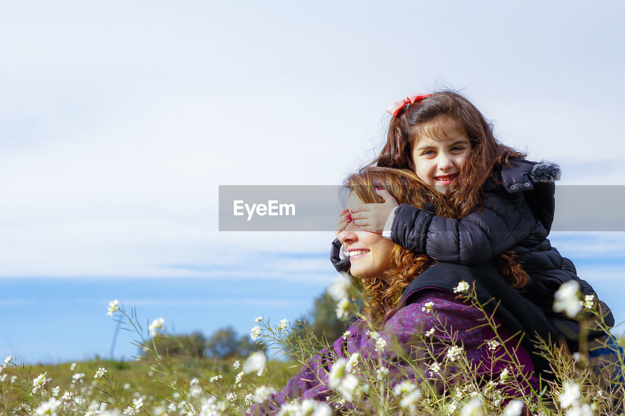 Mother with daughter sitting on grassy field amidst flowers against sky