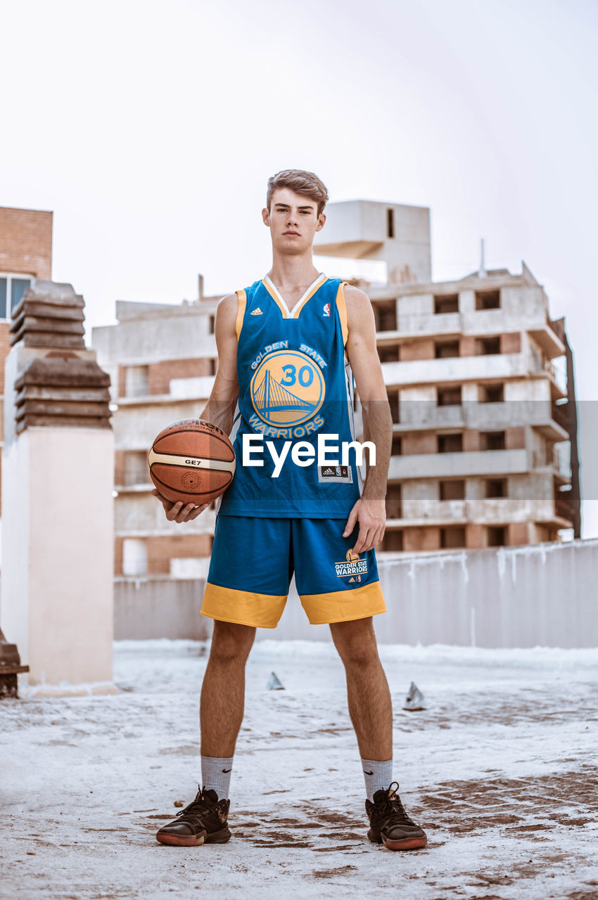 Portrait of young man holding basketball while standing against building