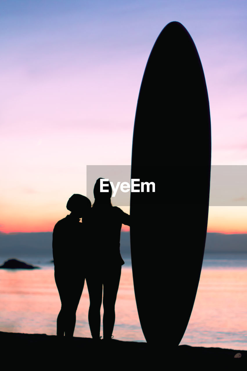 Silhouettes of two friends and surfboard at the seashore during sunset