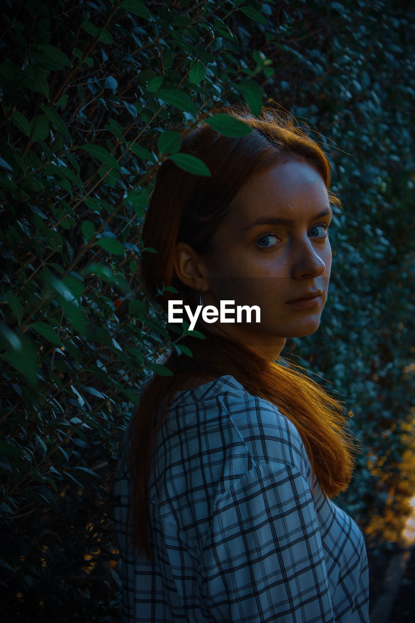 PORTRAIT OF YOUNG WOMAN LOOKING AWAY WHILE STANDING BY PLANTS