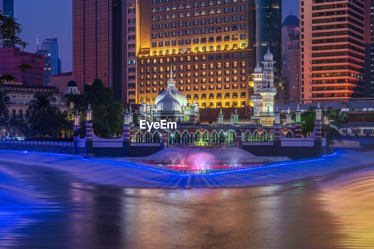 ILLUMINATED FOUNTAIN IN CITY BY BUILDING AT NIGHT