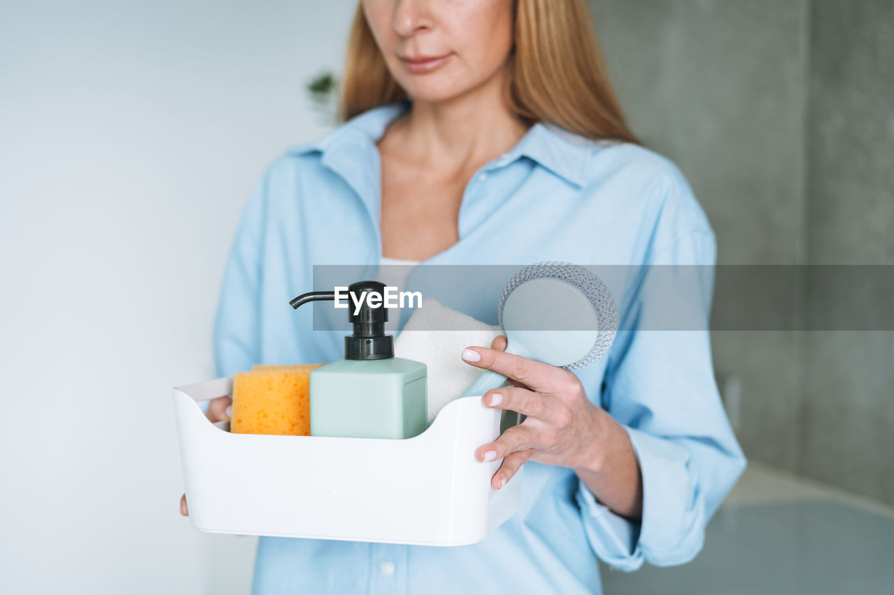 Young woman in blue shirt holding box of things for cleaning the kitchen at home