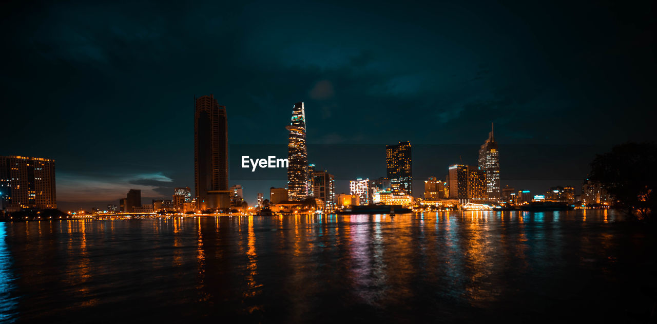 Illuminated buildings by river against sky at night