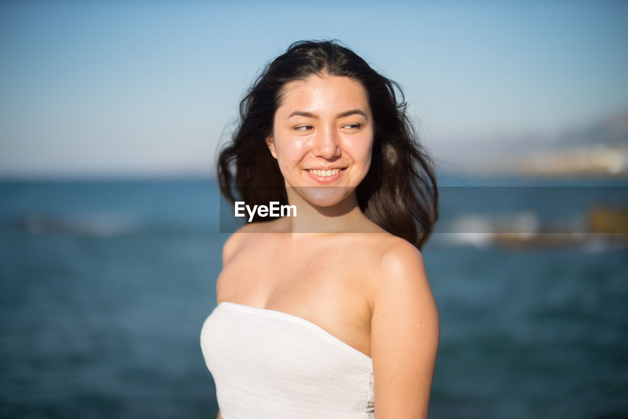 Smiling young woman looking away while standing against sea on sunny day