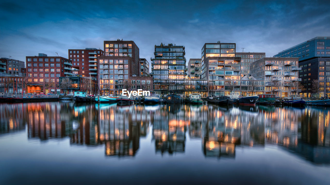 ILLUMINATED BUILDINGS BY RIVER AGAINST SKY AT DUSK