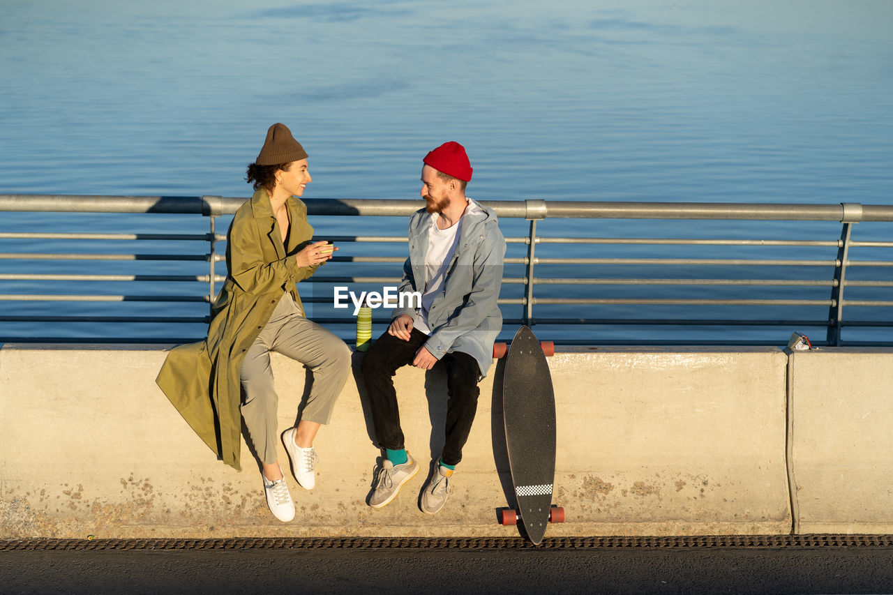 Young couple of hipsters enjoy time together sit on bridge at river drink hot tea from thermos talk