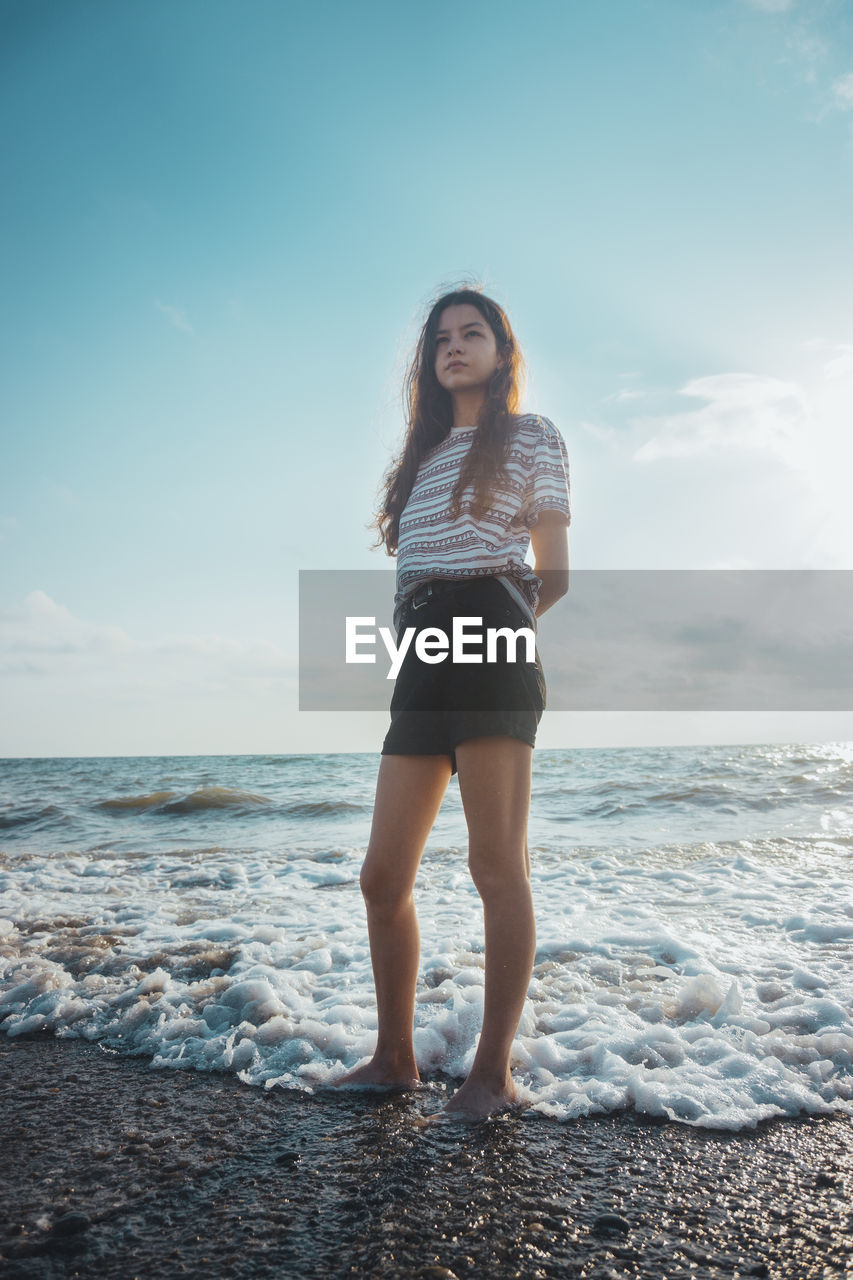 FULL LENGTH OF YOUNG WOMAN STANDING ON BEACH AGAINST SKY