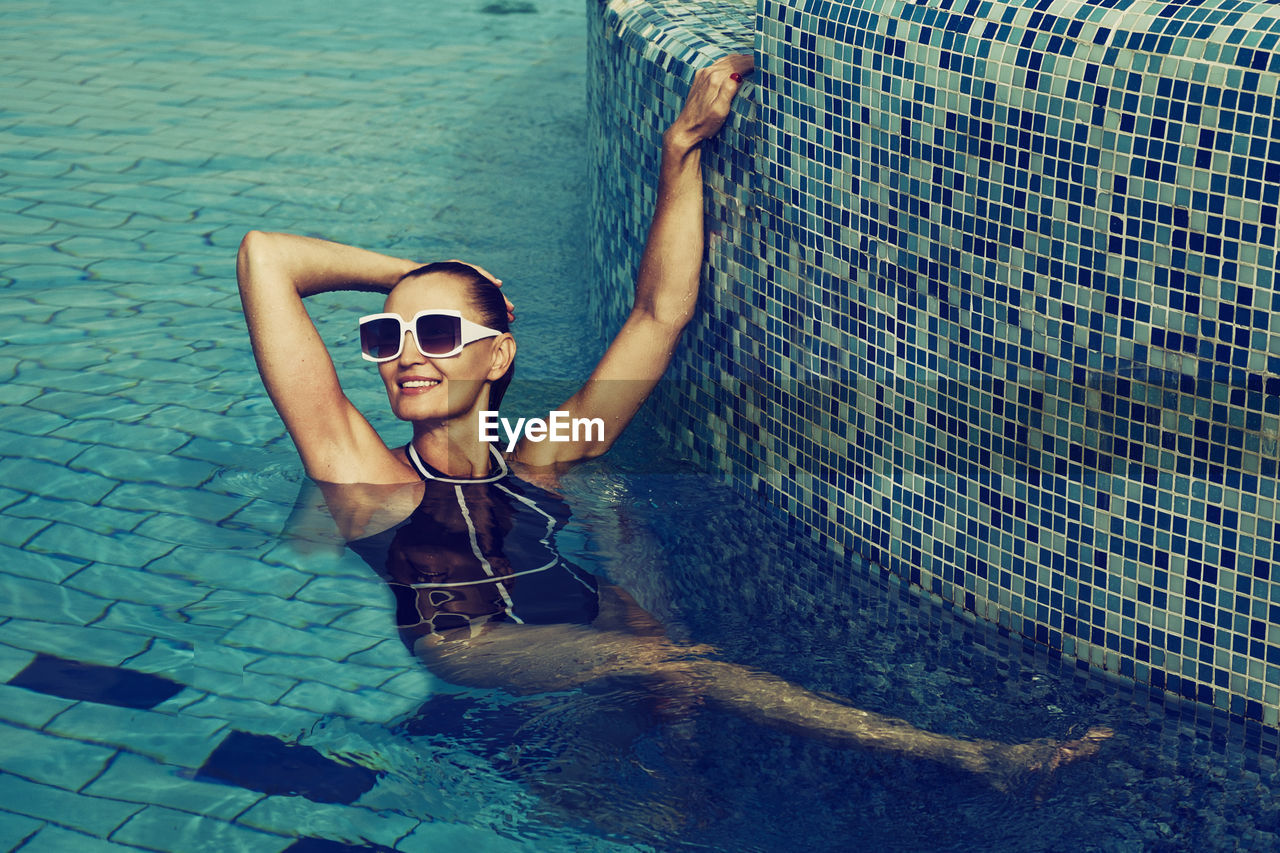 A vintage photo of a woman in a black swimsuit and white sunglasses swimming in the pool