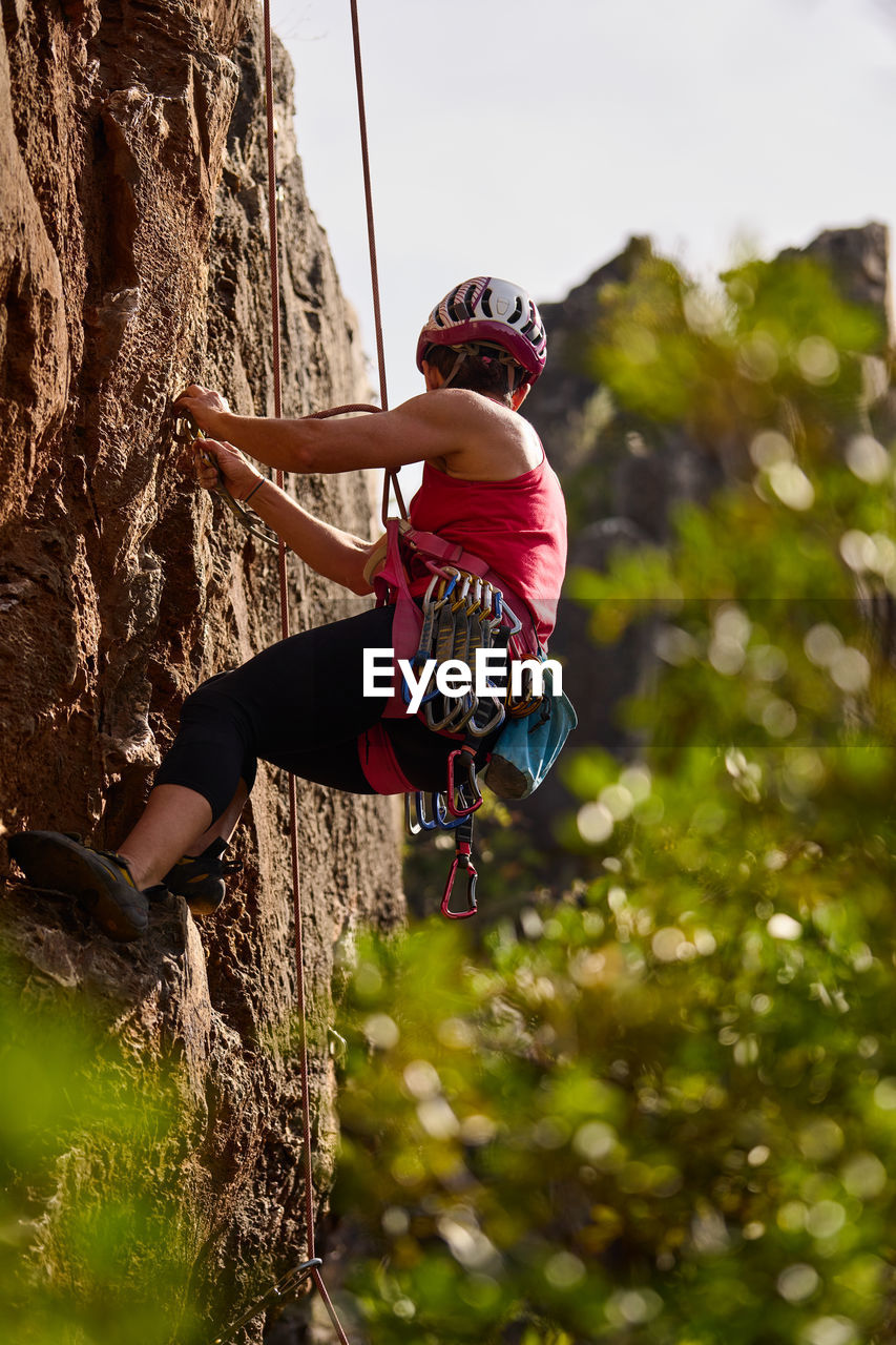 Low angle full length of active senior woman climbing on rocky cliff against sky