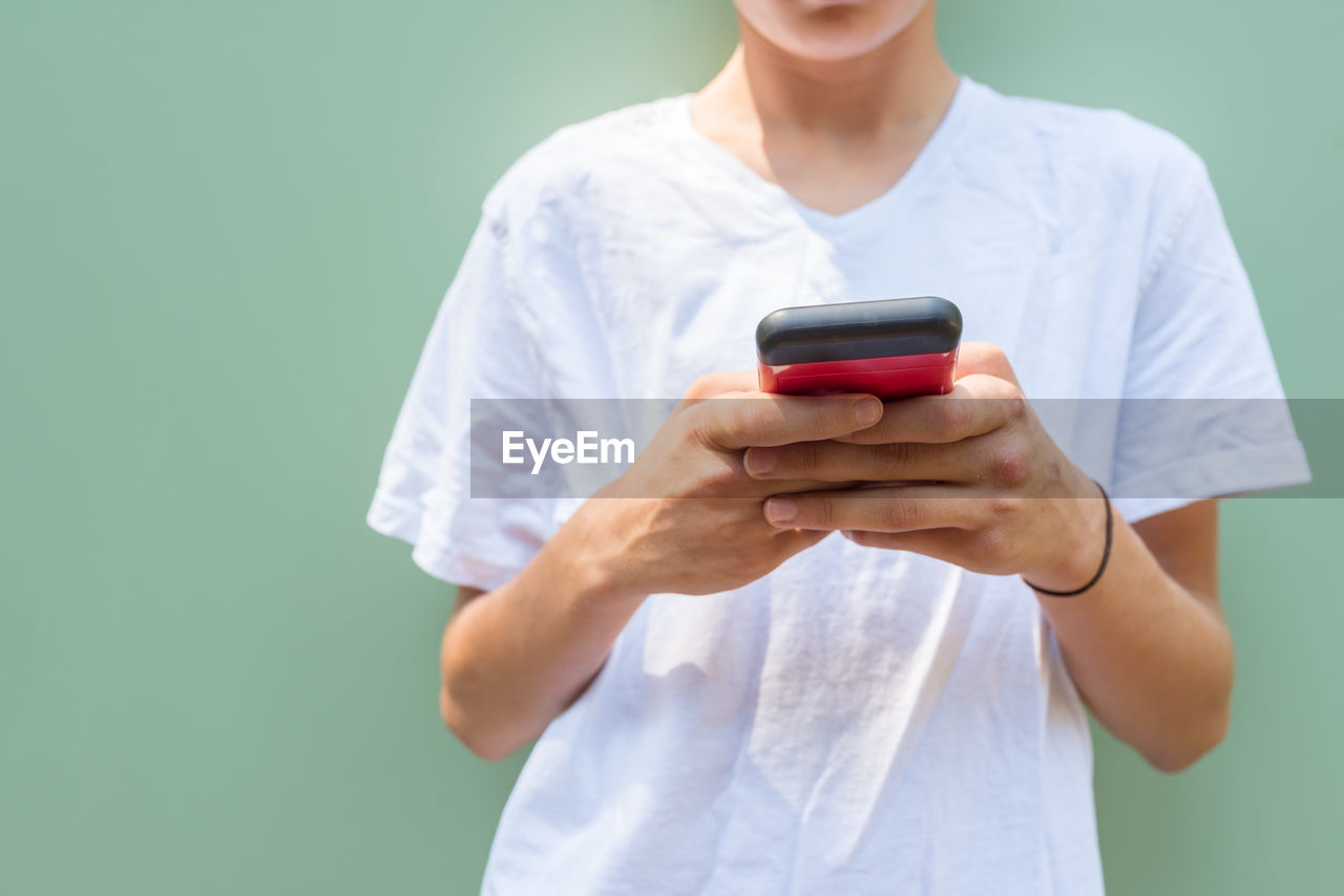 Midsection of teenage boy using mobile phone while standing against wall