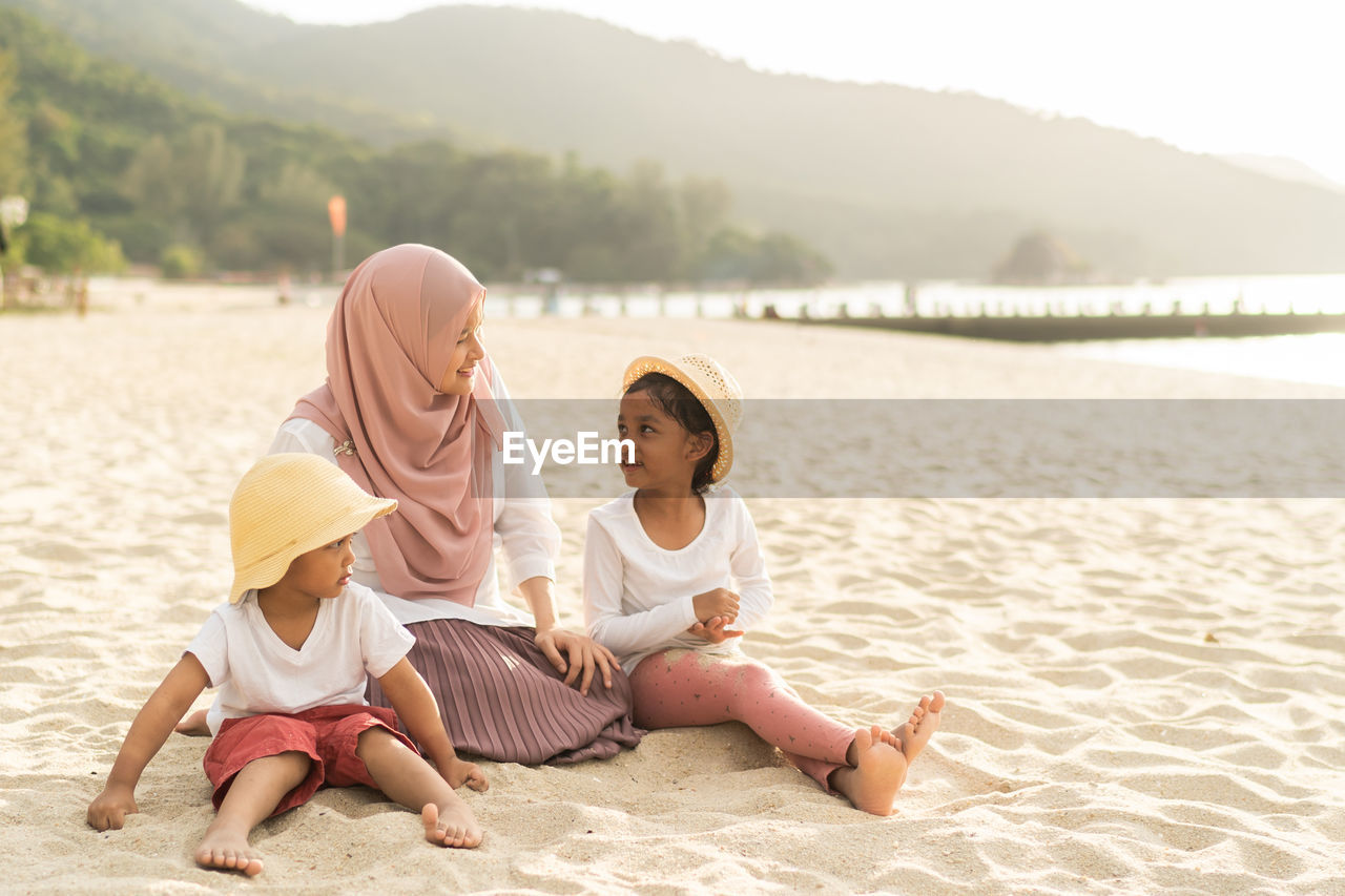 Mother wearing hijab with children sitting on sand at beach